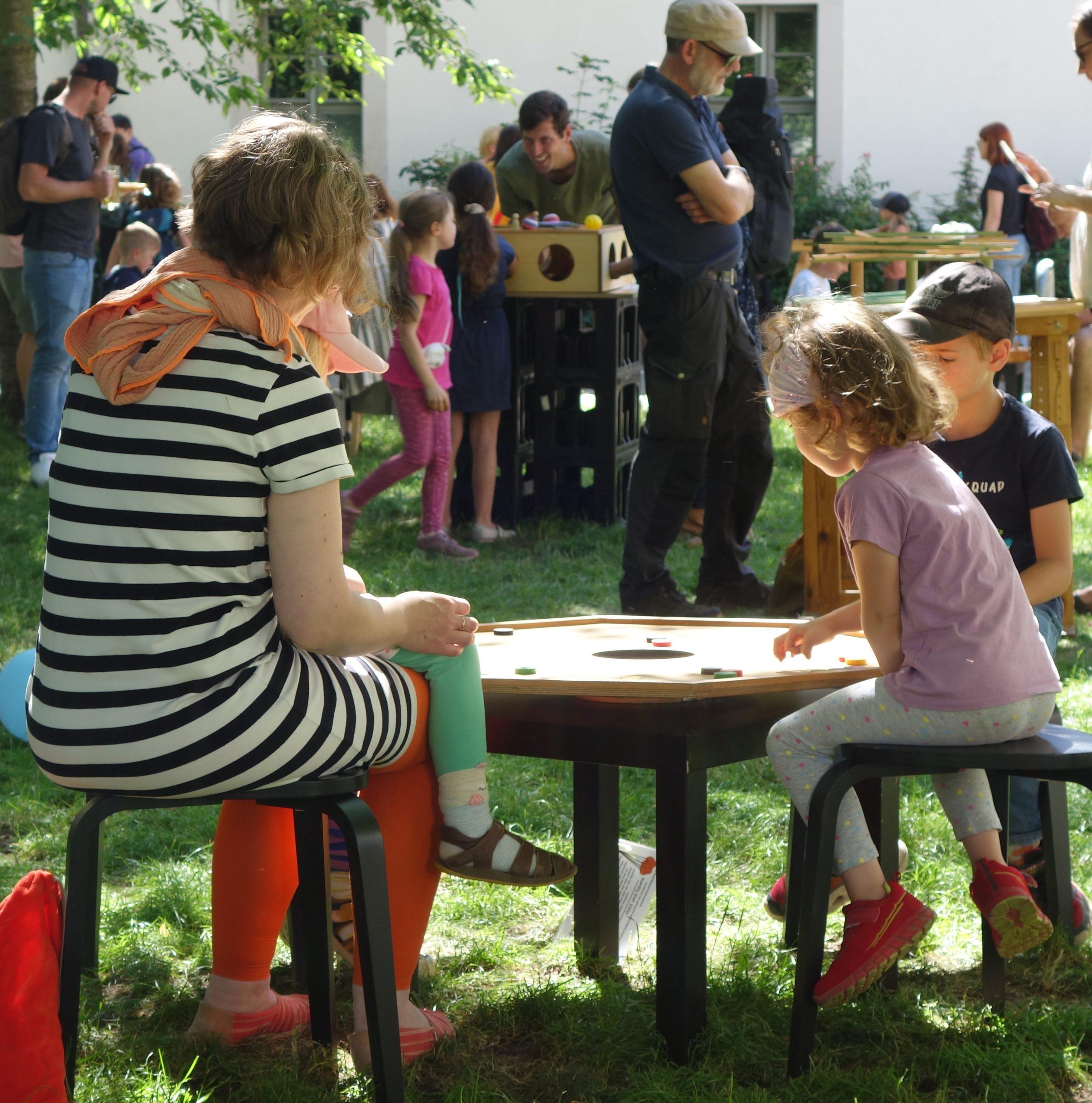 Kinder und Erwachsene spielen ein Brettspiel an einem Tisch. Im Hintergrund sind weitere spielende Menschen zu sehen.